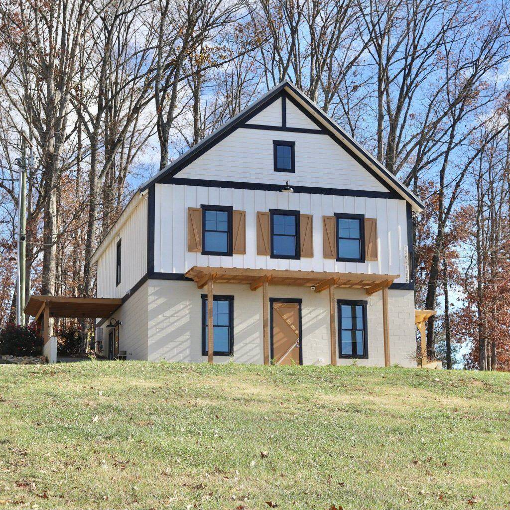 A modern farmhouse with white siding and black trim sits on a grassy hillside surrounded by bare trees.