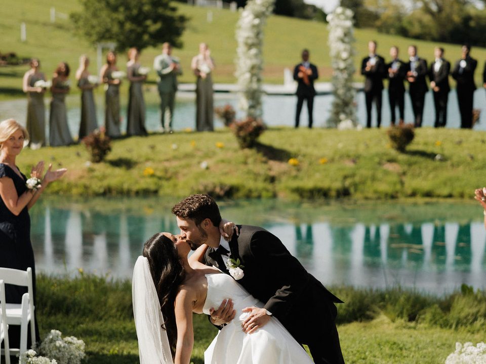 Un novio inclina a su novia en un beso sobre la isla del estanque, con el cortejo alineado junto al agua en Cedar Pond Farms cerca de Sevierville