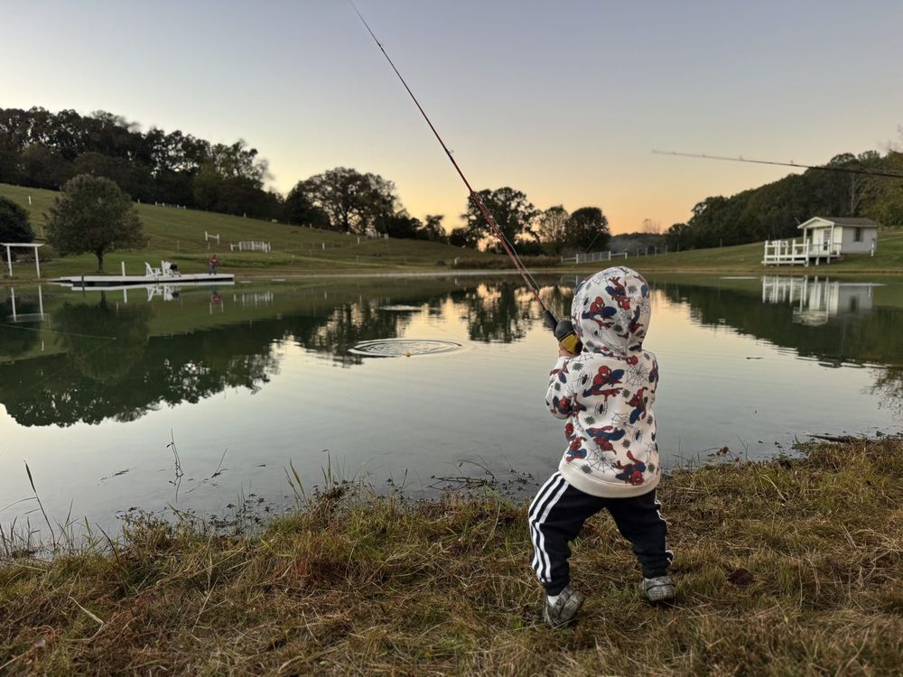 A child casting a fishing rod from the bank of the farm pond at dusk